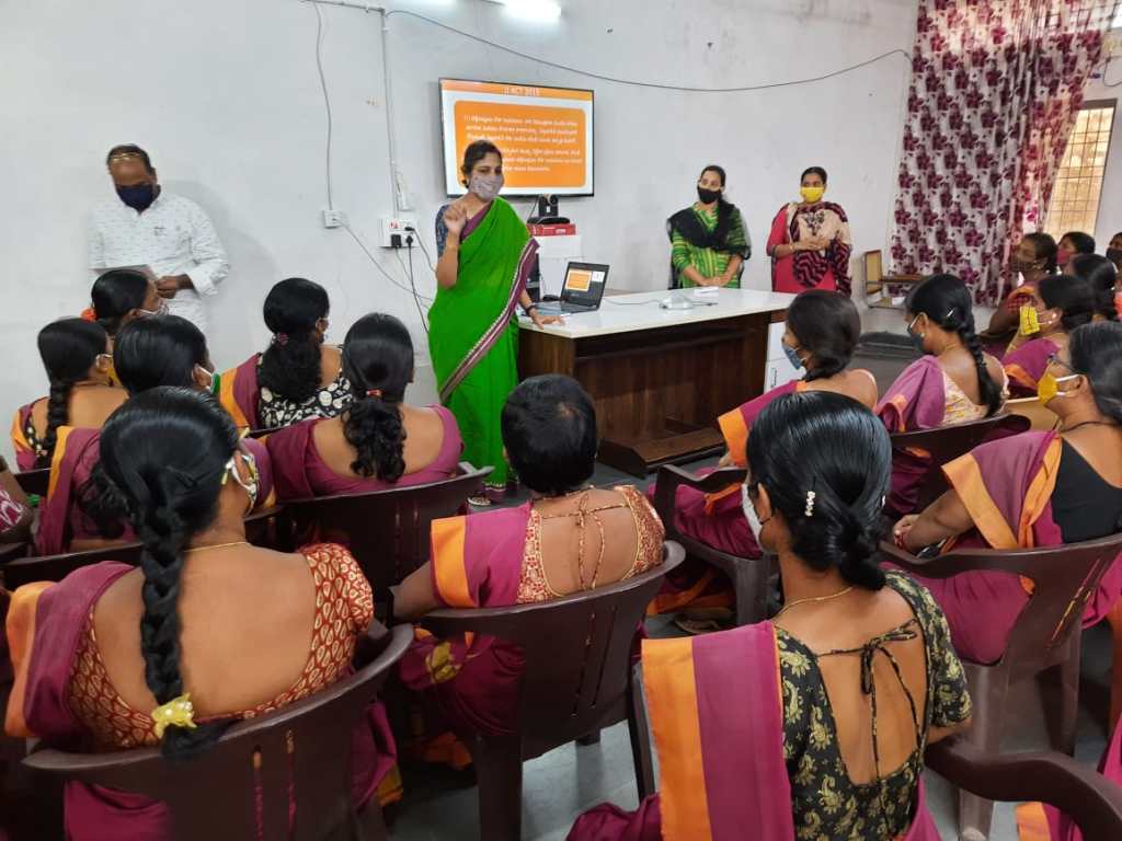 You can see the backs of women in pretty blouses and saris, and a woman in green sari in the front. She is explaining things, there is a laptop and on the wall, at TV, on which the contents of the slides from the laptop are shared